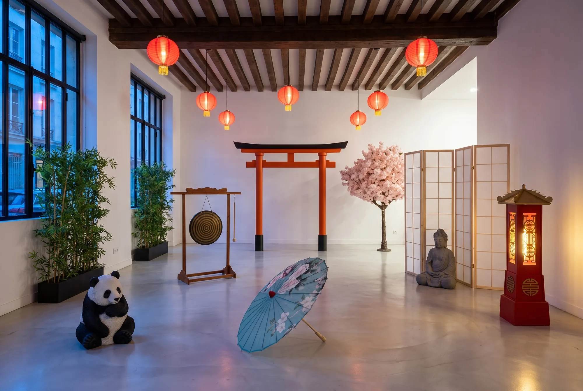 Espace de cocktail d'inspiration japonaise dans une salle aux poutres apparentes : arche asiatique rouge, cerisier en fleurs, paravent japonais, statue de Bouddha, gong sur son support en bois, lanterne XL rouge et dorée, bambous en jardinière, statue de panda, parasol asiatique ouvert et lampions chinois suspendus