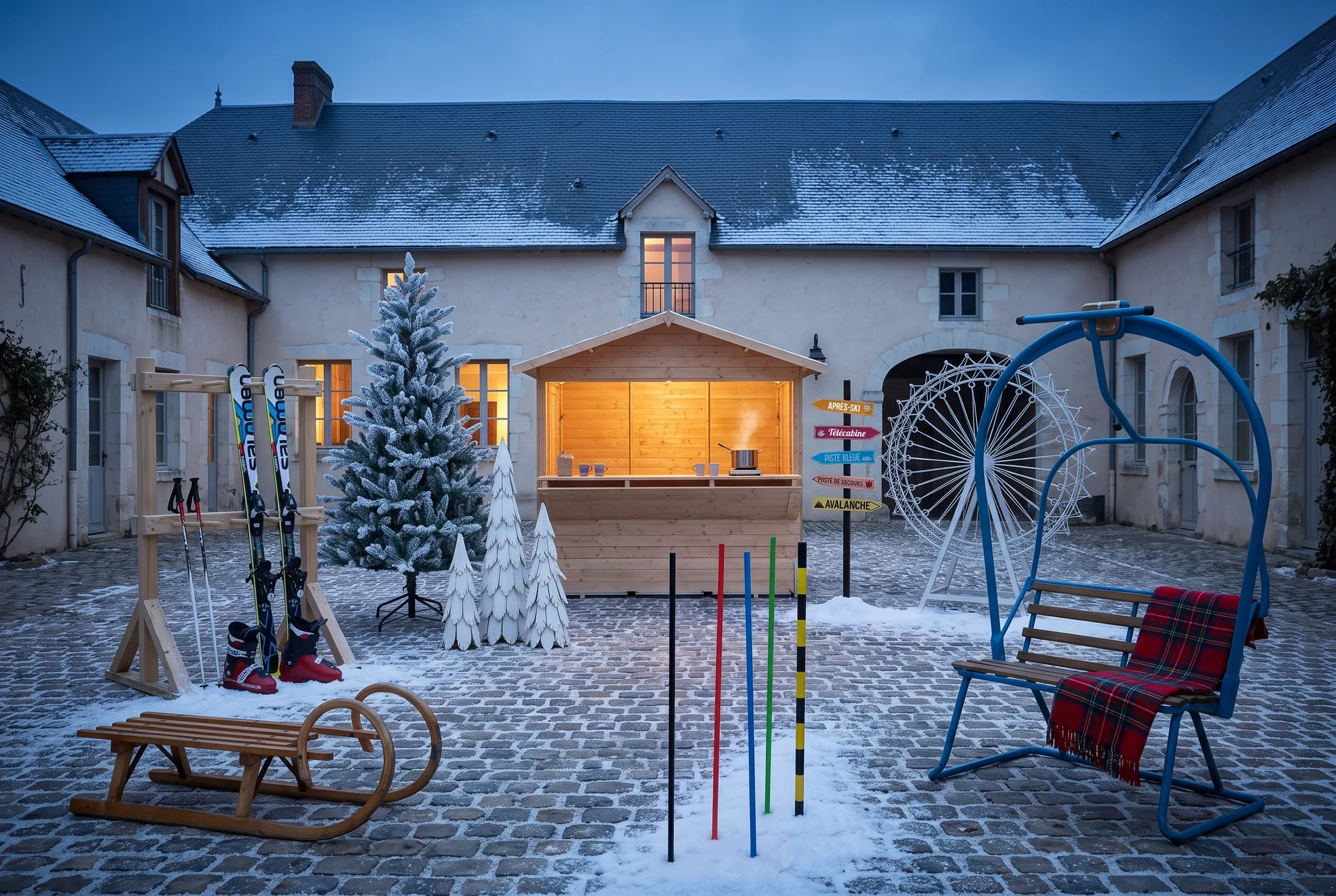 Village d'hiver dans la cour pavée et enneigée d'un domaine à la tombée de la nuit : chalet démontable en bois éclairé, sapin enneigé, lot de trois sapins en bois blanc, grande roue blanche, télésiège bleu, rack à skis avec skis, bâtons et chaussures, luge en bois, panneau directionnel et jalons de signalisation de ski