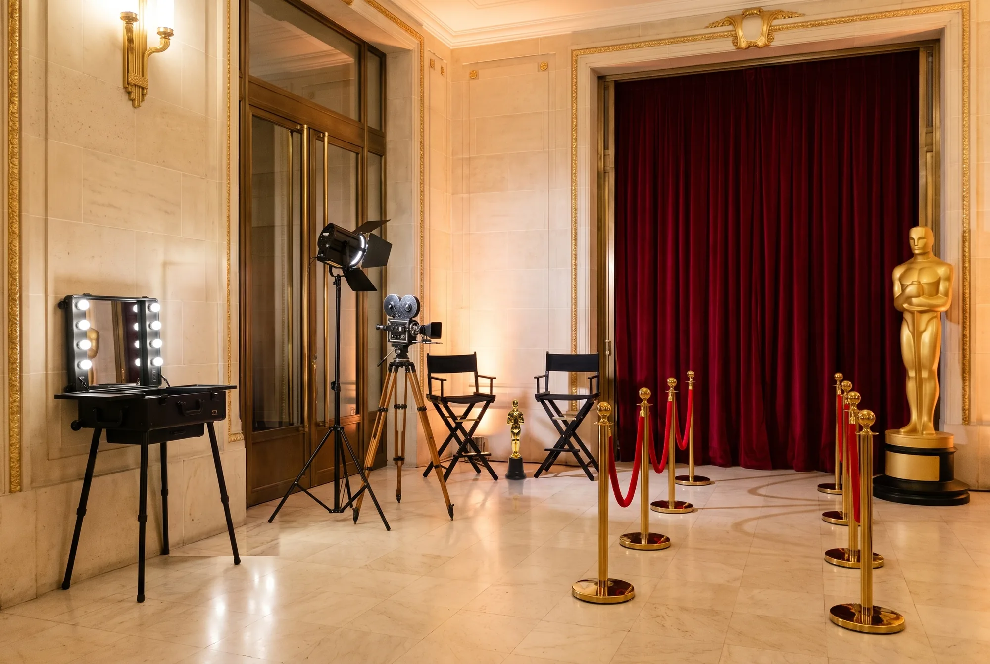 Soirée de gala à thème cinéma dans le foyer d'une salle de réception : rideau de velours rouge, allée de potelets dorés à cordon rouge, grande statue dorée, statuette dorée sur socle noir, deux chaises hautes de réalisateur, caméra ancienne sur trépied, projecteur de studio et table de maquillage lumineuse