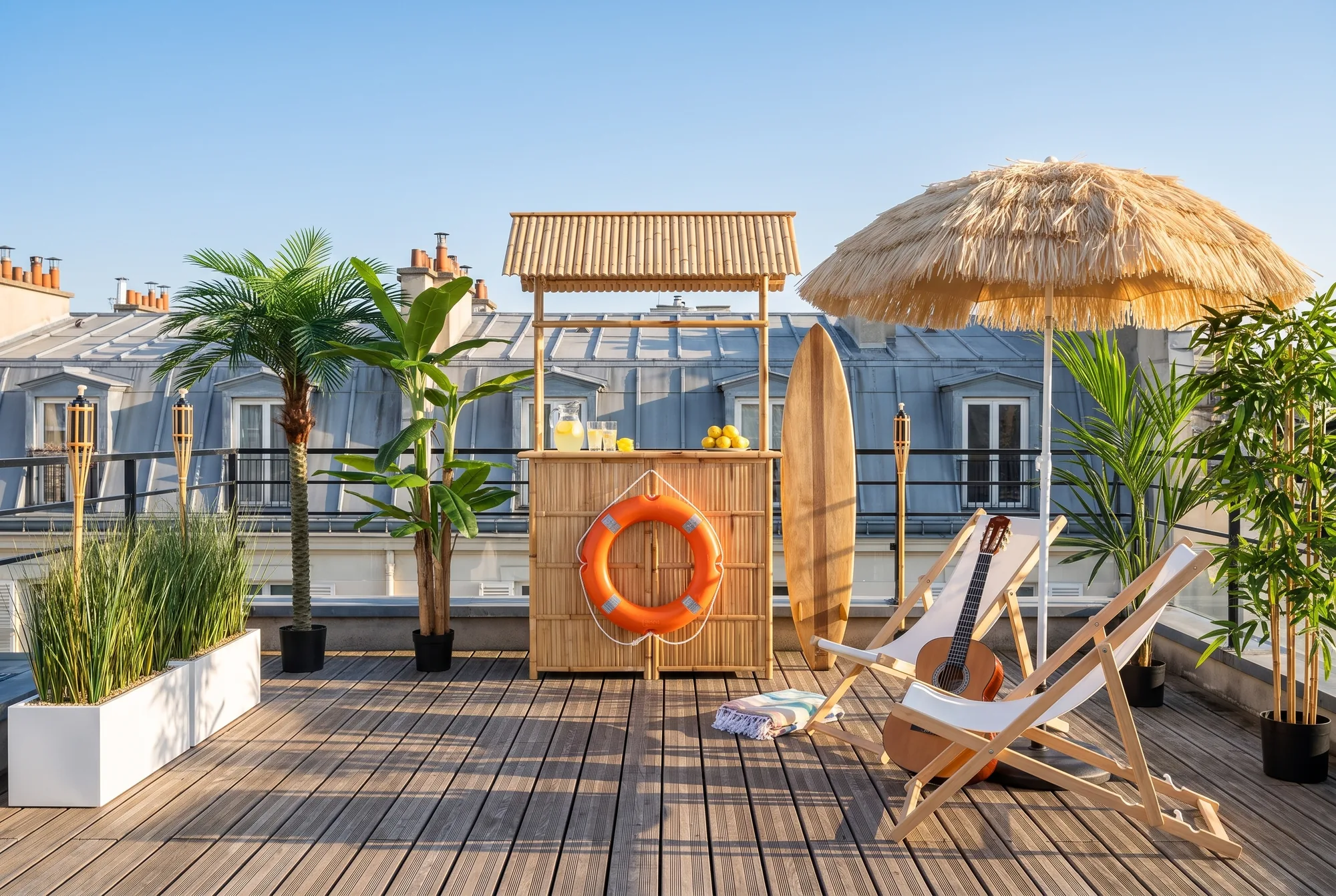 Terrasse sur les toits de Paris en bar de plage : bar en bambou avec bouée de sauvetage, planche de surf, deux transats et une guitare sous un parasol en paille, palmier, bananier, kentia, bambous, jardinières d'herbes hautes et torches en bambou