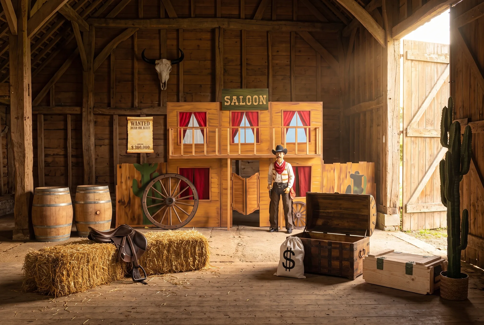 Grange aménagée en décor western : façade de saloon en bois, statue de cowboy, tonneaux, bottes de paille et selle, roue de calèche, affiche Wanted, crâne de bison, coffre en bois et sac de dollars, caisse de munitions, cactus en pot