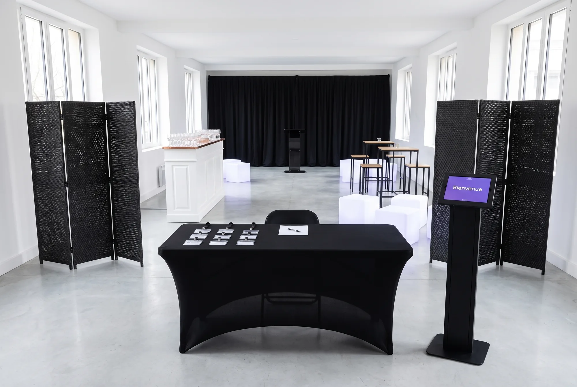 Salle de réception d'entreprise : table d'accueil habillée d'une nappe en lycra noire, borne tablette affichant « Bienvenue », deux paravents noirs, bar blanc, tables hautes et tabourets, cubes lumineux, rideaux noirs et pupitre de conférence