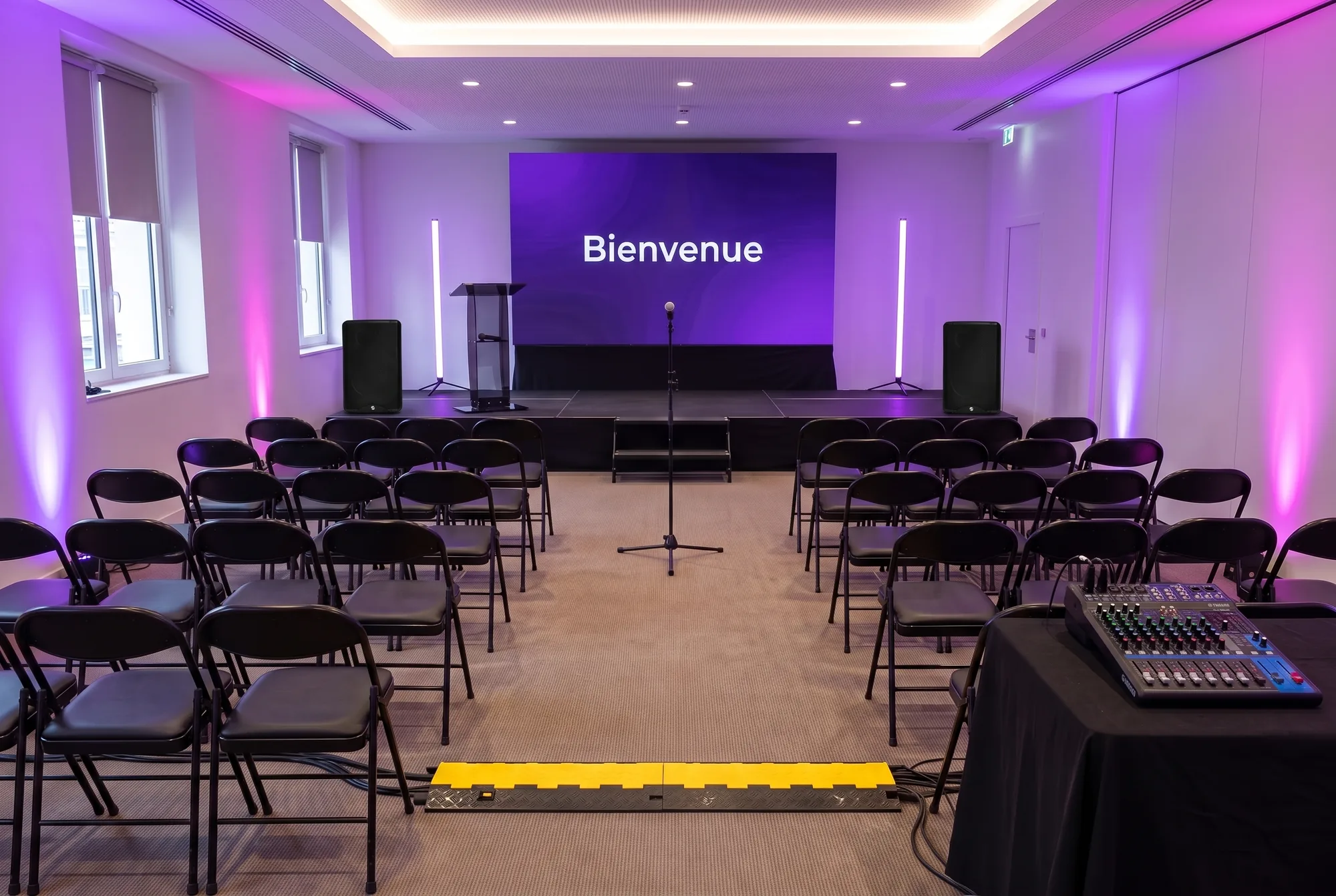 Salle de conférence préparée : scène basse, écran LED affichant « Bienvenue », pupitre, enceintes, tubes LED, éclairage violet des murs, rangées de chaises, micro sur pied dans l'allée et console de mixage