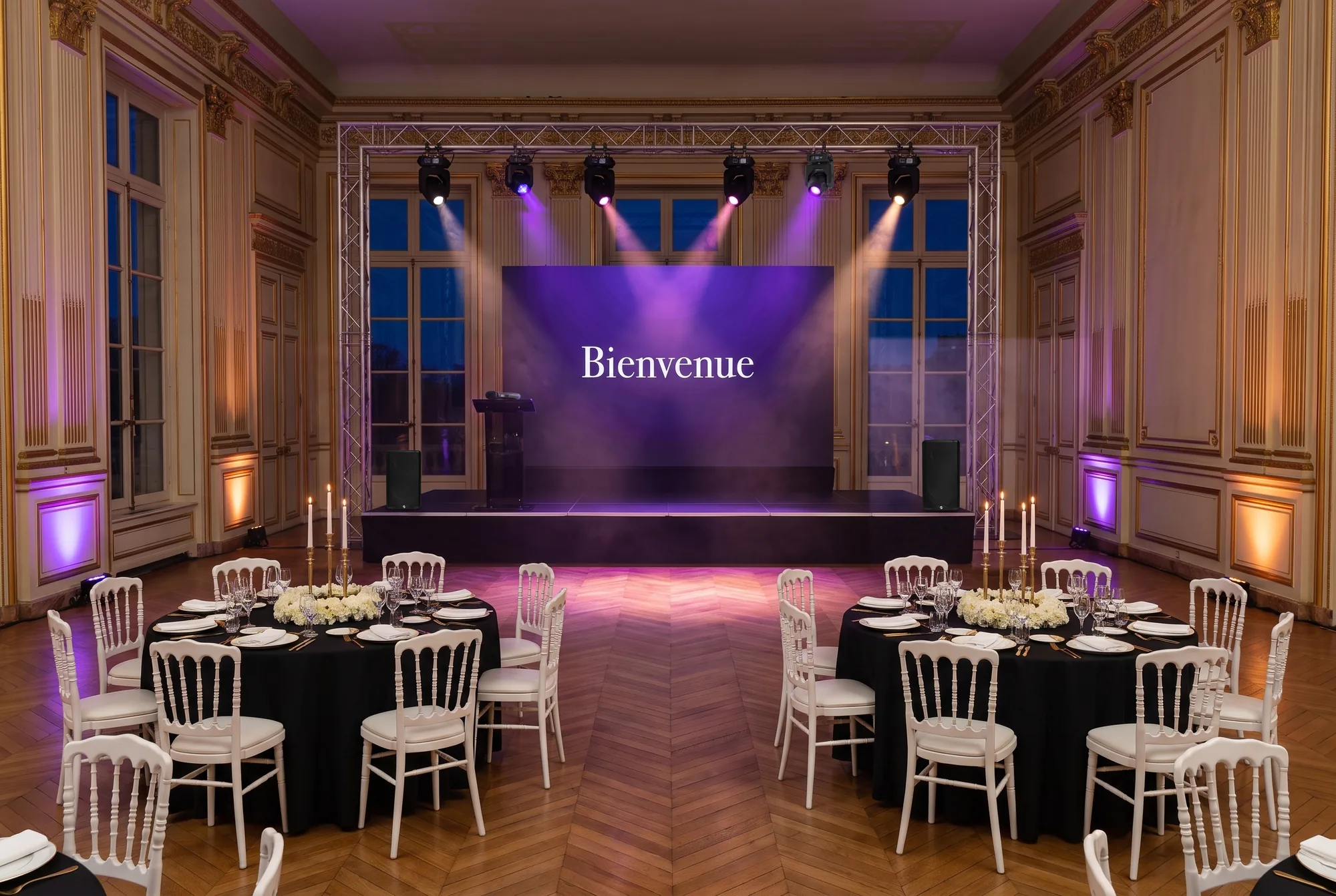 Salon de réception haussmannien préparé pour la soirée annuelle d'une entreprise : tables rondes nappées de noir, chaises blanches Napoléon III, chandeliers dorés, scène en praticables avec écran LED affichant « Bienvenue », pupitre et micro sans fil, enceintes, portique en truss et lyres au-dessus de la piste