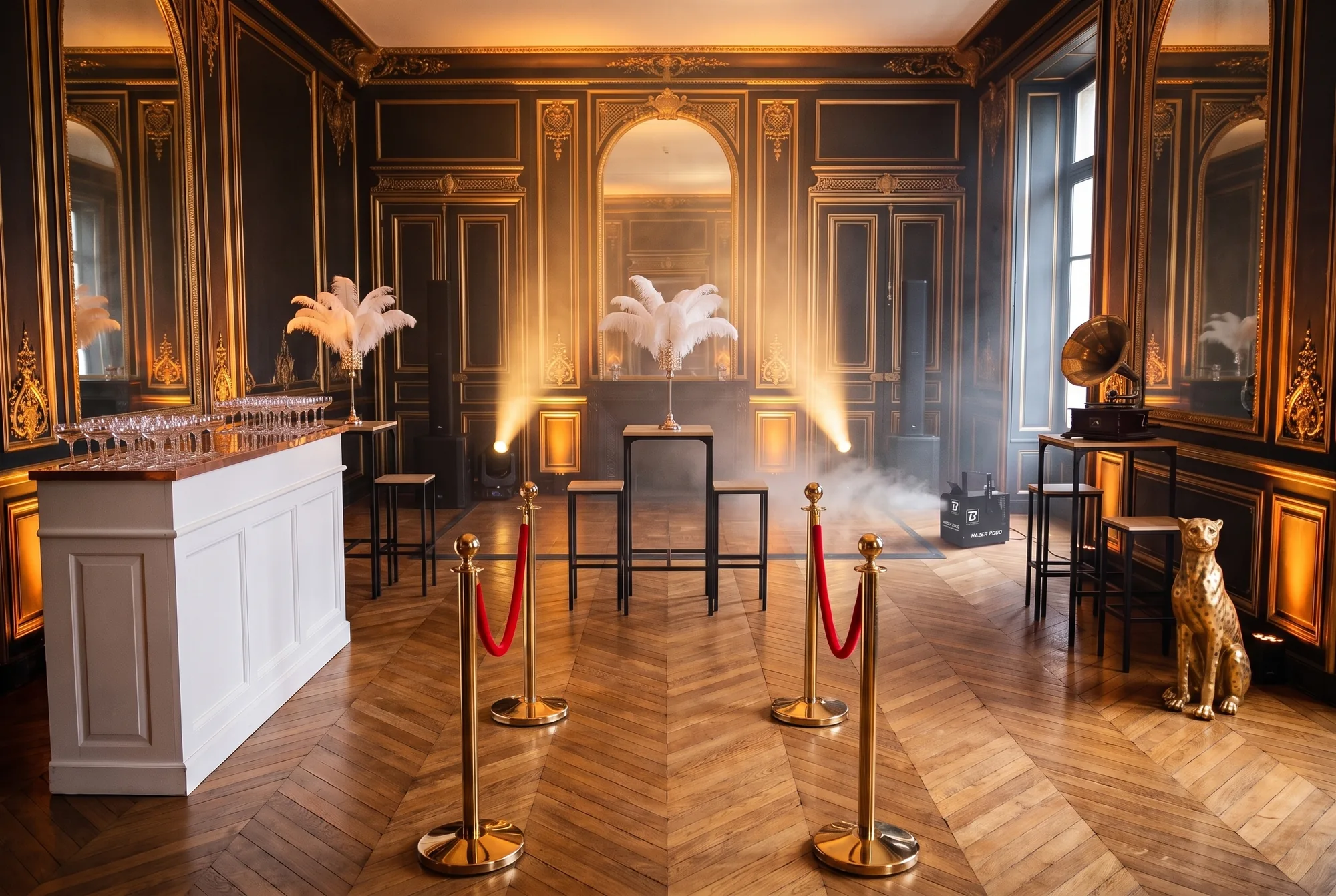 Salon d'hôtel particulier préparé pour une soirée Gatsby : potelets dorés à cordons rouges, bar blanc chargé de coupes, tables hautes et tabourets, centres de table en plumes d'autruche, gramophone, statue de guépard dorée, enceintes colonnes sur caissons, lyres et machine à brume
