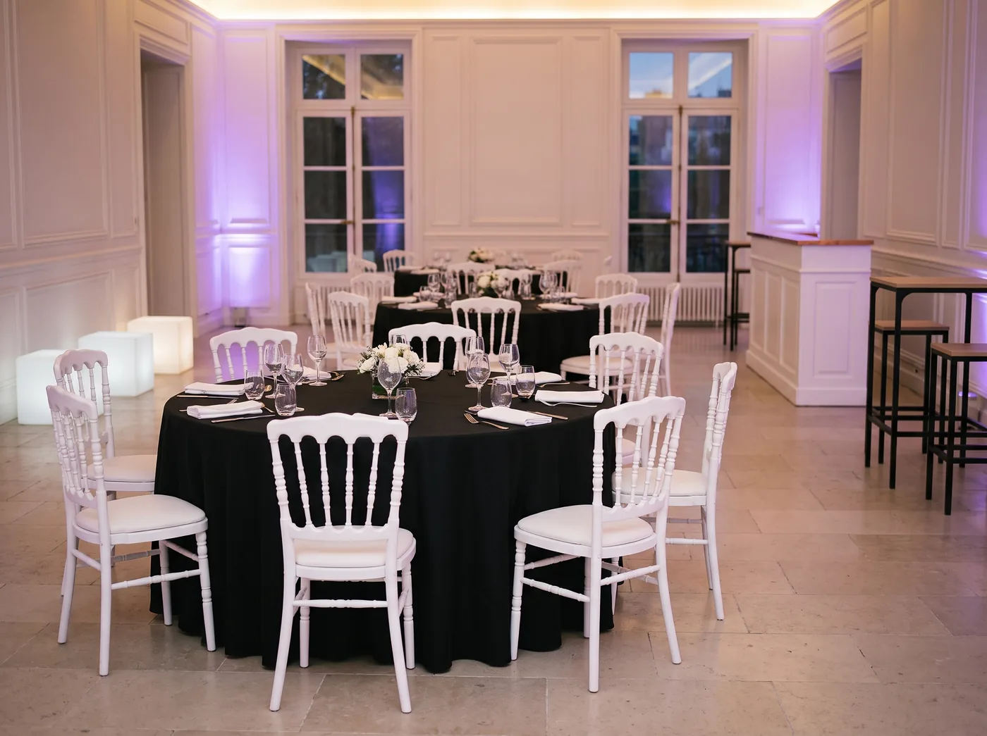 Salle de réception dressée : tables rondes en nappes noires, chaises Napoléon III blanches, bar et tables hautes